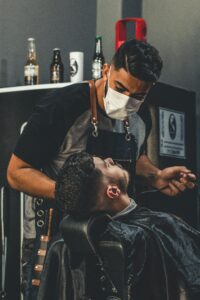 A barber wearing a face mask gives a haircut to a client in an indoor barbershop setting.