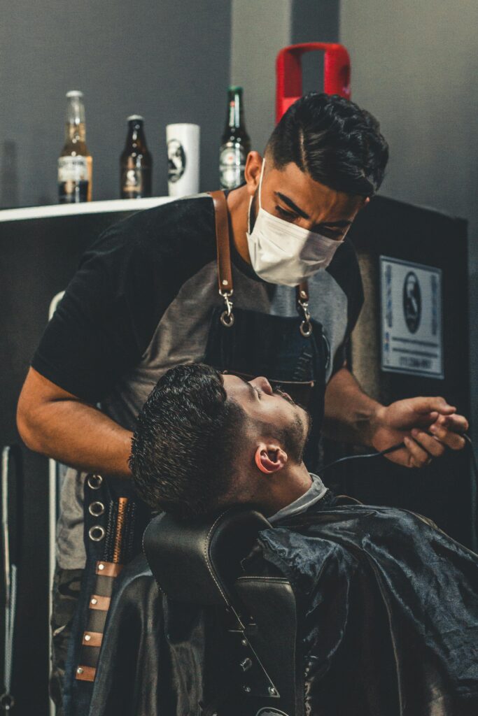 A barber wearing a face mask gives a haircut to a client in an indoor barbershop setting.